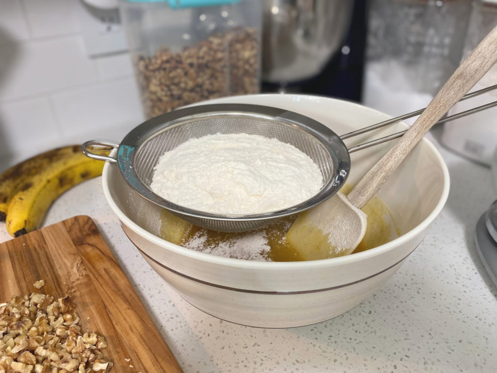 A wire mesh strainer filled with flour sitting on a bowl with a spatula. [decadent banana bread]