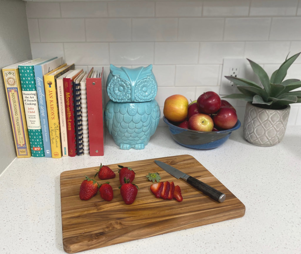 A cutting board on a counter with sliced strawberries, and cookbooks and a cookie jar in the background. [Strawberry Shortcake]