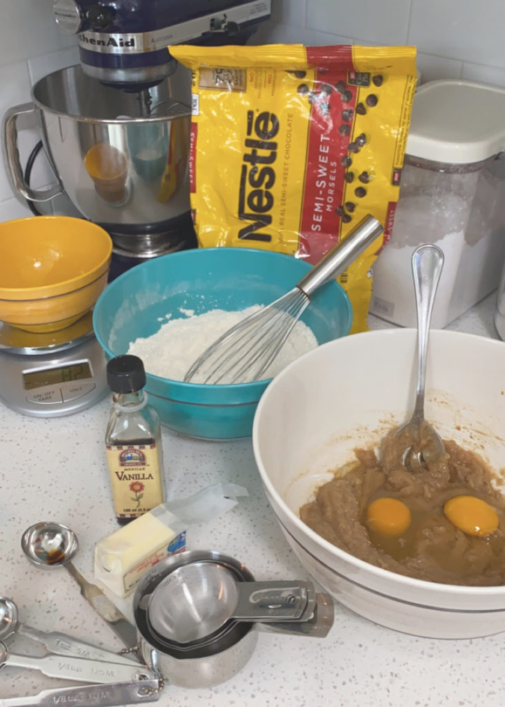 A bowl with sugar, eggs, and butter with other ingredients on the counter. [chocolate chip cookie bars]