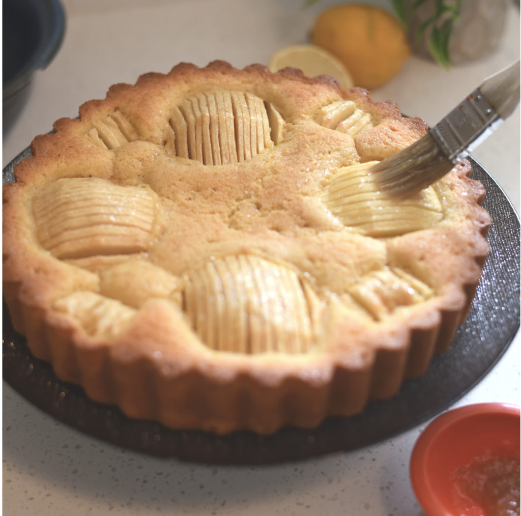 German sunken apple cake on a cake plate being brushed with orange marmalade. [sunken apple cake]