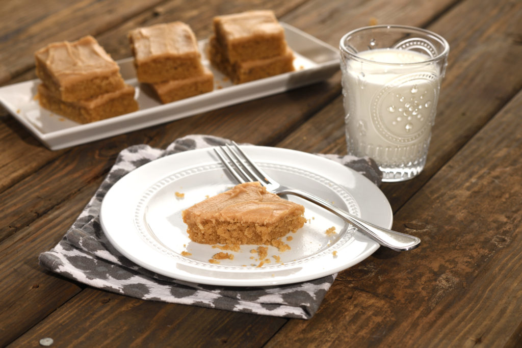 A slice of peanut butter sheet cake with peanut butter fudge frosting on a plate with a glass of milk