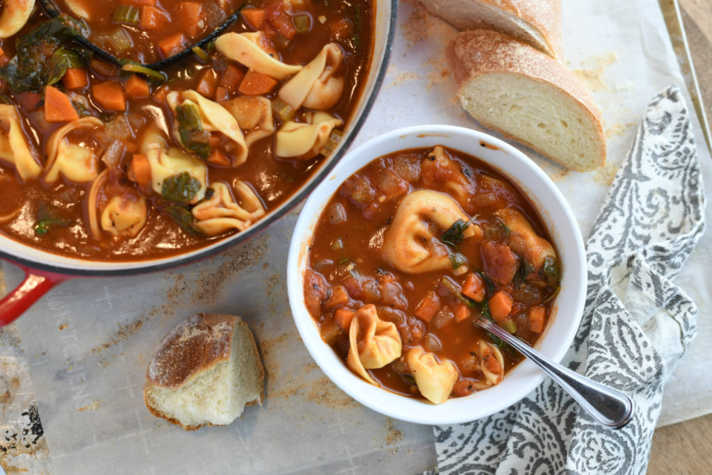 A pot of tortellini soup with a small bowl f soup and a loaf of french bread.