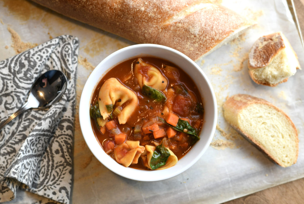 A bowl of tortellini soup with french bread in background.