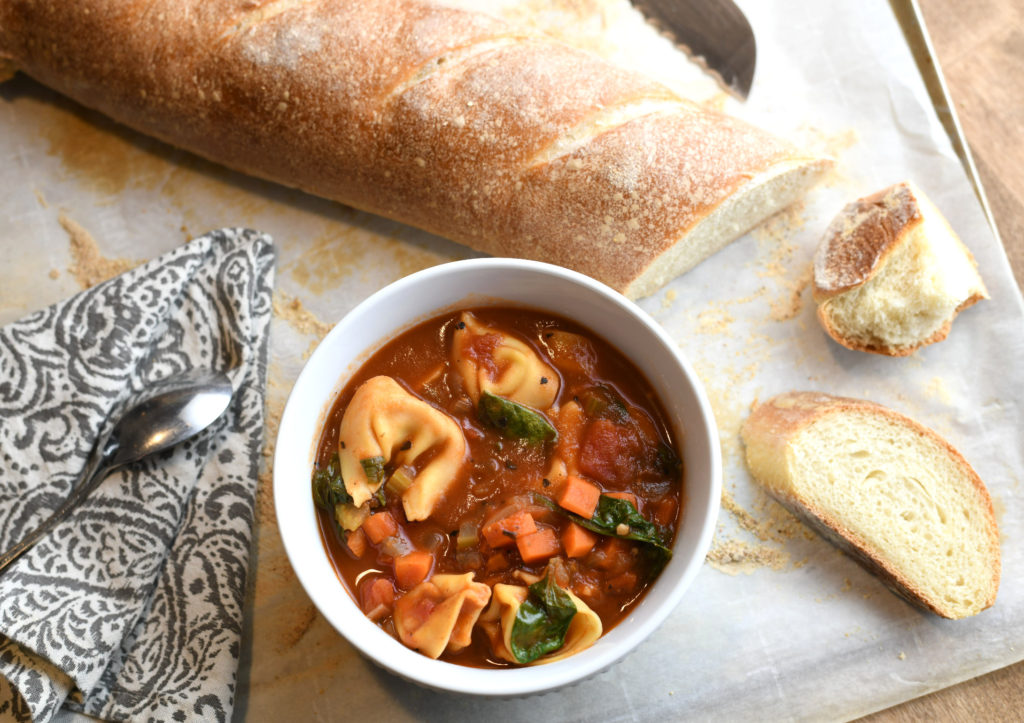 A bowl of tortellini soup with a sliced loaf of french bread.